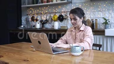 年轻女子专心致志地坐在厨房的木桌旁，用她的<strong>银色笔记本</strong>电脑和一杯